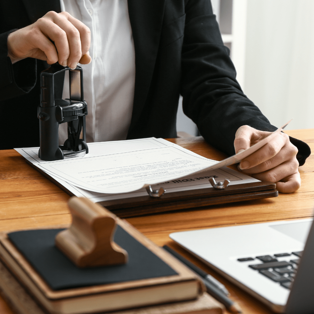 A close-up shot of a Notary Public in a black blazer using a stamp on a legal document on a wooden desk. A clipboard, a laptop, books, and a wooden stamper are visible on the table.